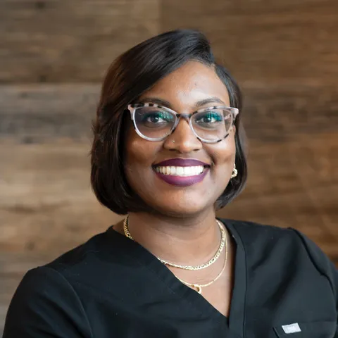 Smiling woman with glasses and short hair wearing black scrubs, gold necklaces.