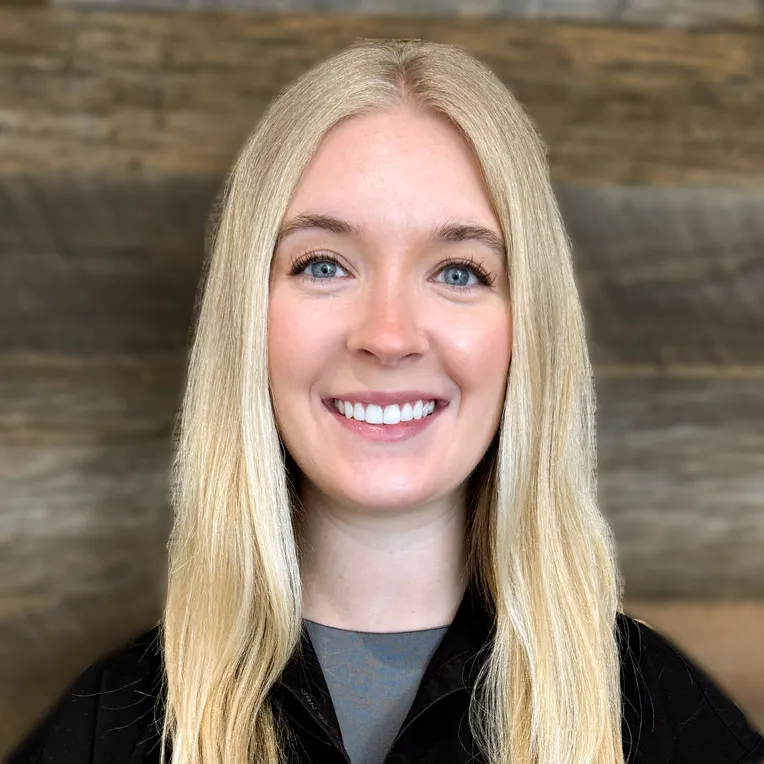 Smiling woman with long blonde hair wearing a black jacket against a wood background.