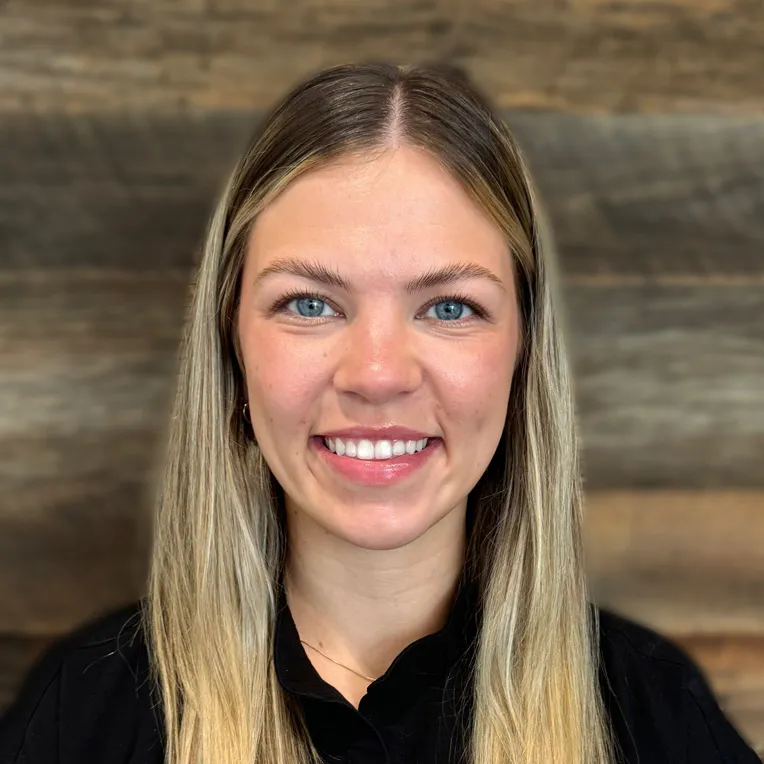 Smiling woman with long blonde hair wearing black collared shirt against wooden background.
