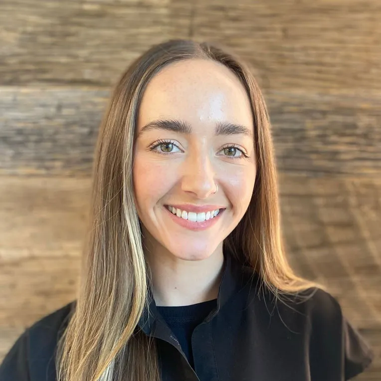 Smiling woman with long straight hair wearing a black jacket against a wooden wall.