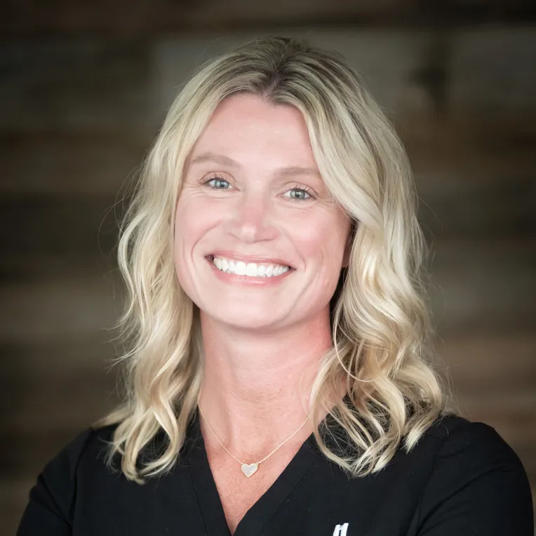 Smiling woman with wavy blonde hair wearing black scrubs and heart necklace.