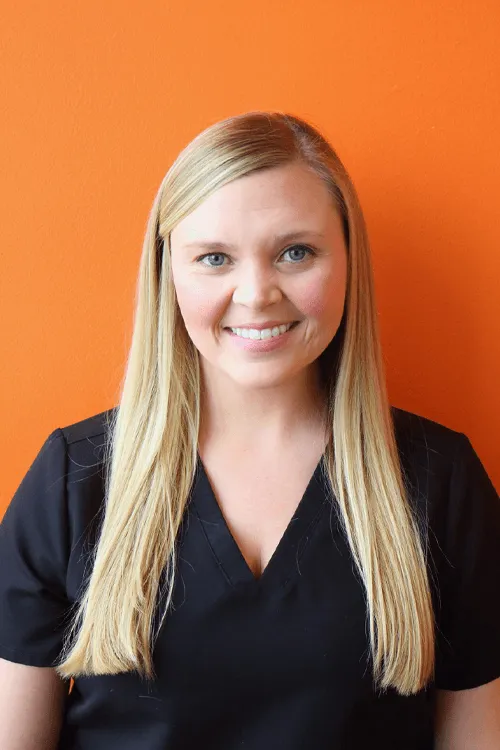 Smiling woman with long blonde hair wearing black scrubs against an orange background.