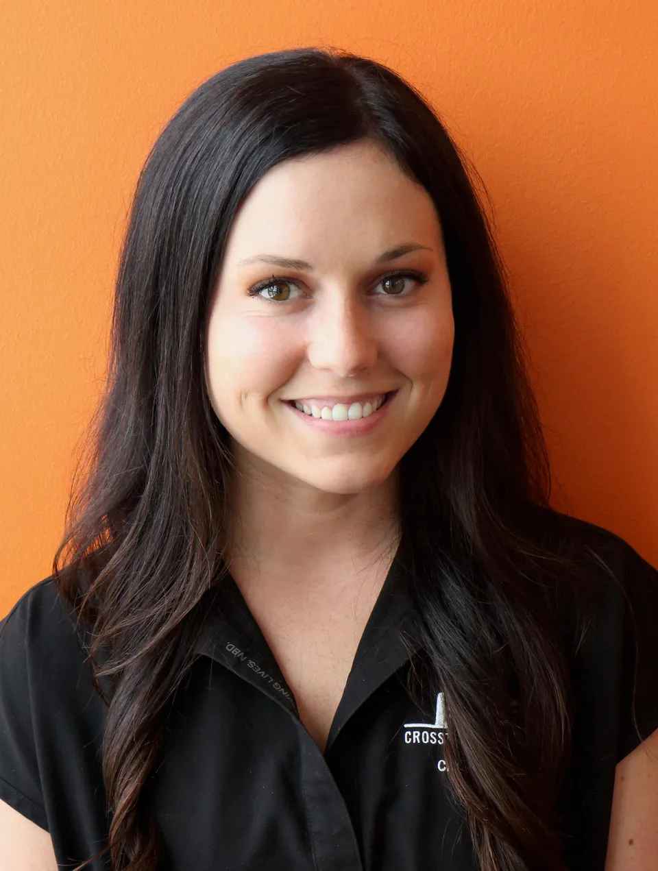 Woman with long dark hair smiling, wearing a black collared shirt with embroidered logo.