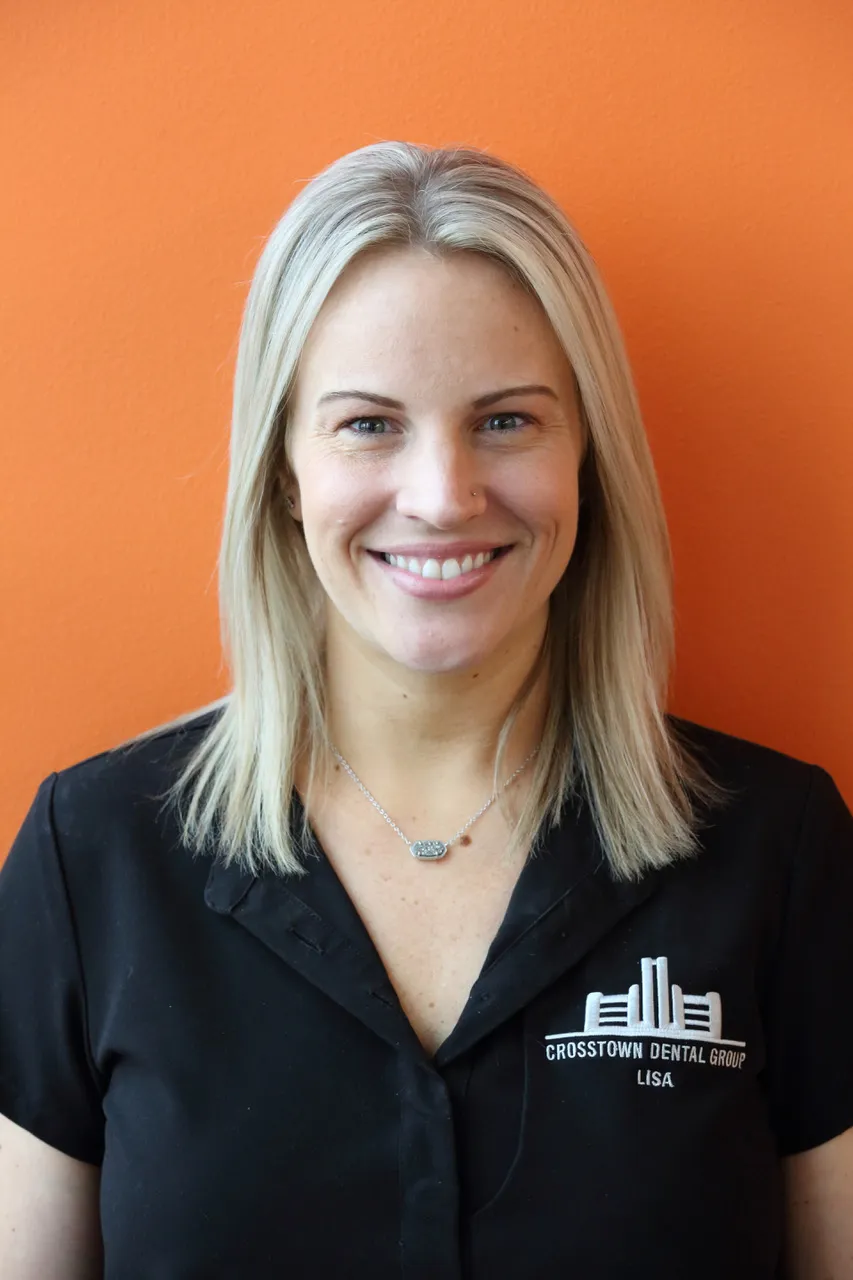 Smiling blonde woman in black Crosstown Dental Group shirt against orange background.